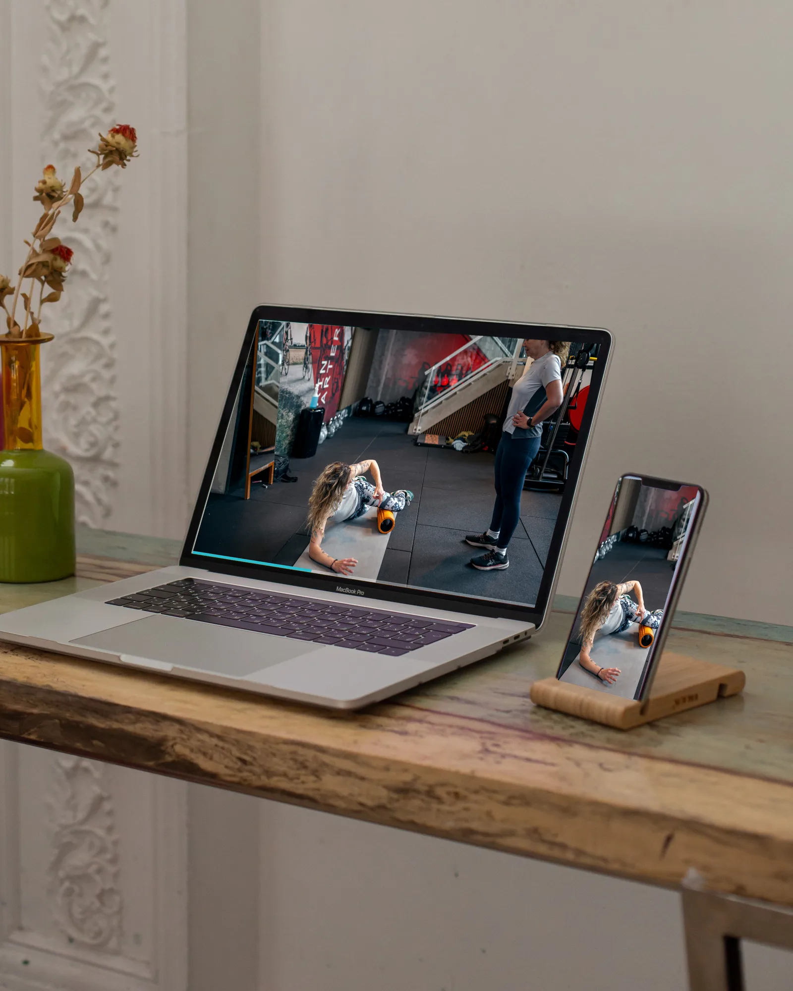 A laptop and a phone on a table at home, both showing Zoë demonstrating an exercise with a foam roller
